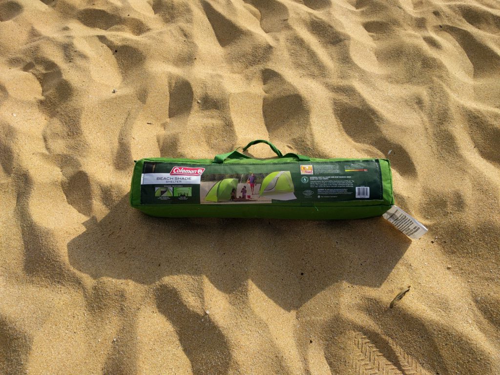 A Coleman Beach Shade carrying case, fully packed, lying on sand.