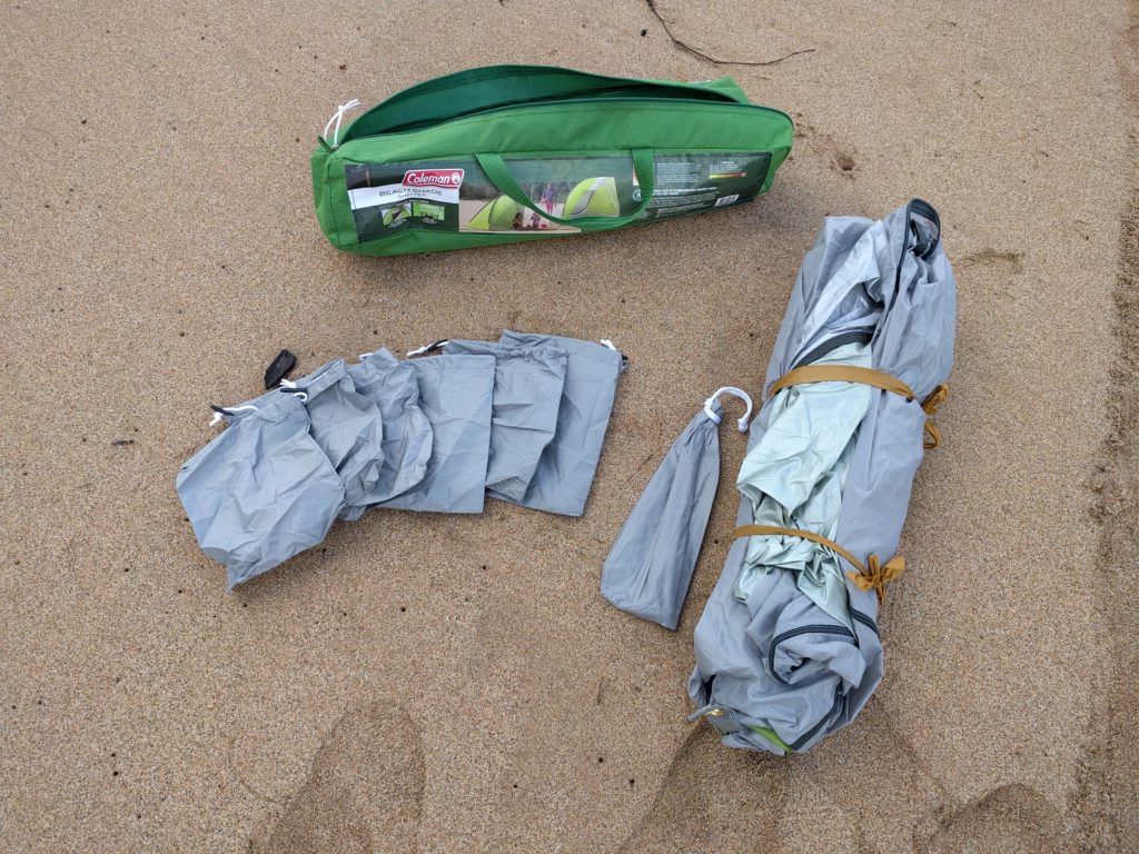 A rolled up Coleman beach tent, its bag of stakes, its sandbags, and its carrying case, laying in the sand.