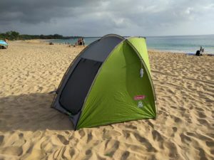 A green Coleman beach tent set up on a sandy beach. The ocean is visible in the background.