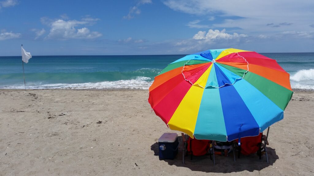A rainbow colored beach umbrella set up on a sandy beach, with the ocean waves visible in the background.